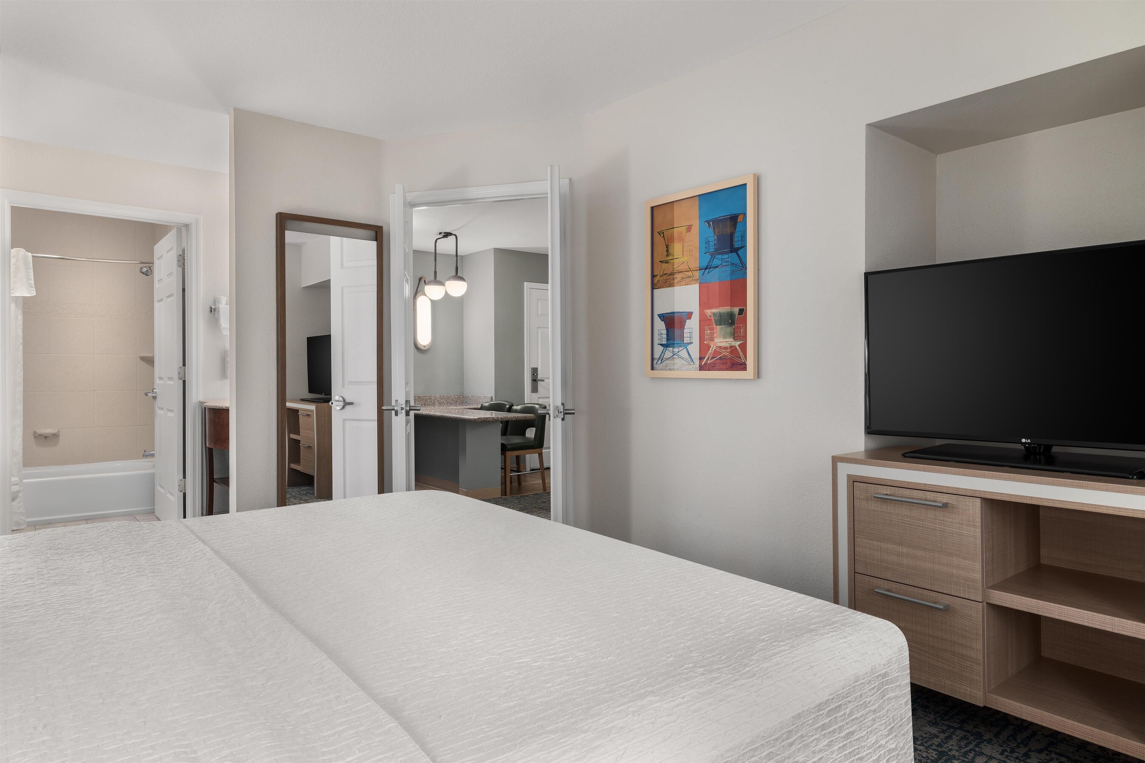 A hotel room with a bed, a TV, a desk, and a bathroom visible through an open door.