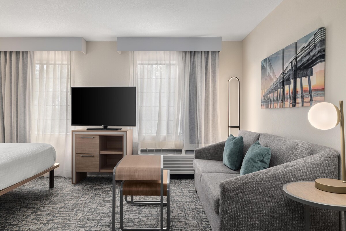 A hotel room with a bed, a sofa, a TV, a desk, and a painting on the wall.