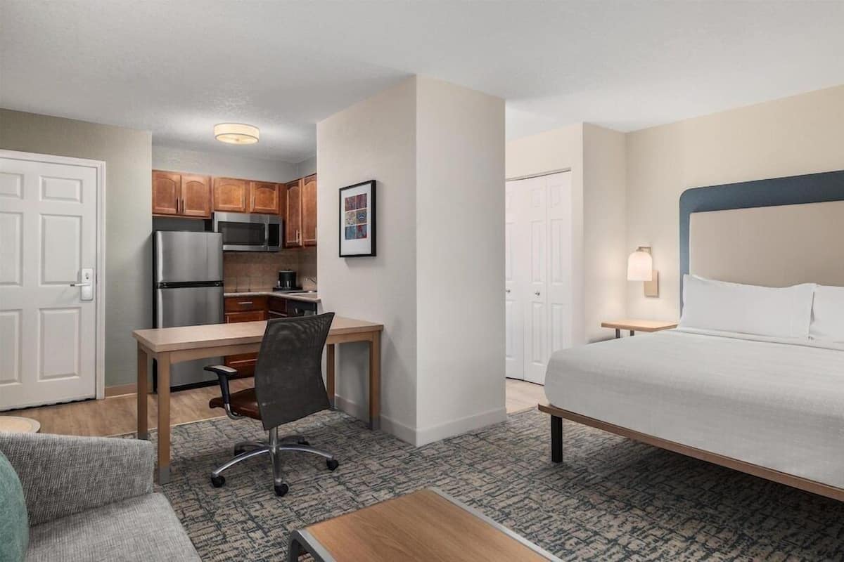 Quarto de hotel pequeno com cama, escrivaninha, cadeira, cozinha compacta e banheiro.