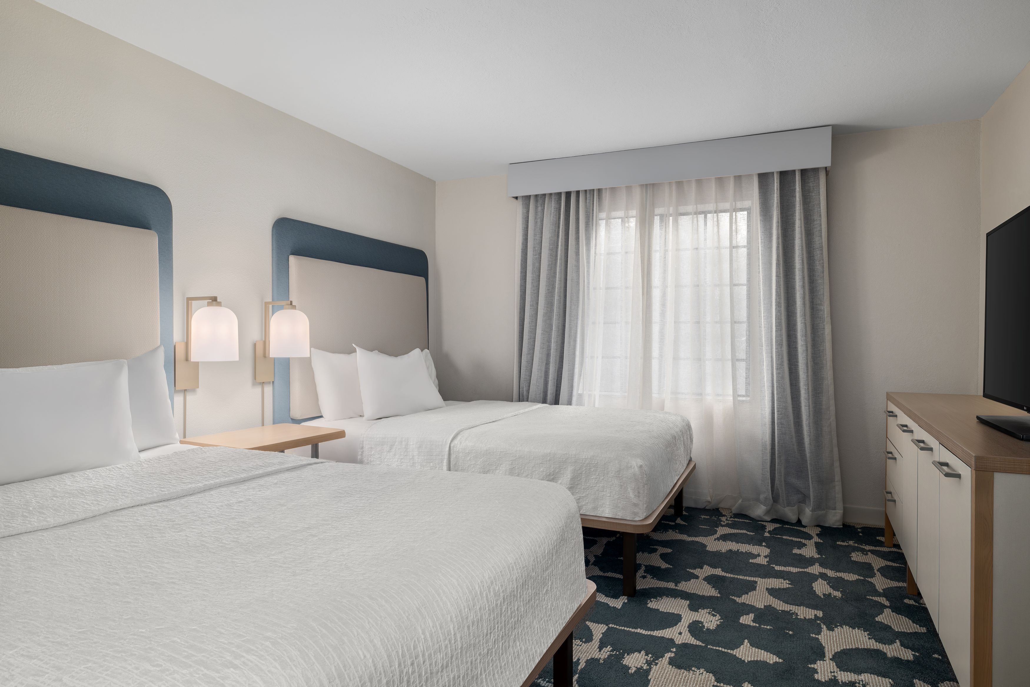 A hotel room with two beds, a desk, a TV, and a window with curtains.