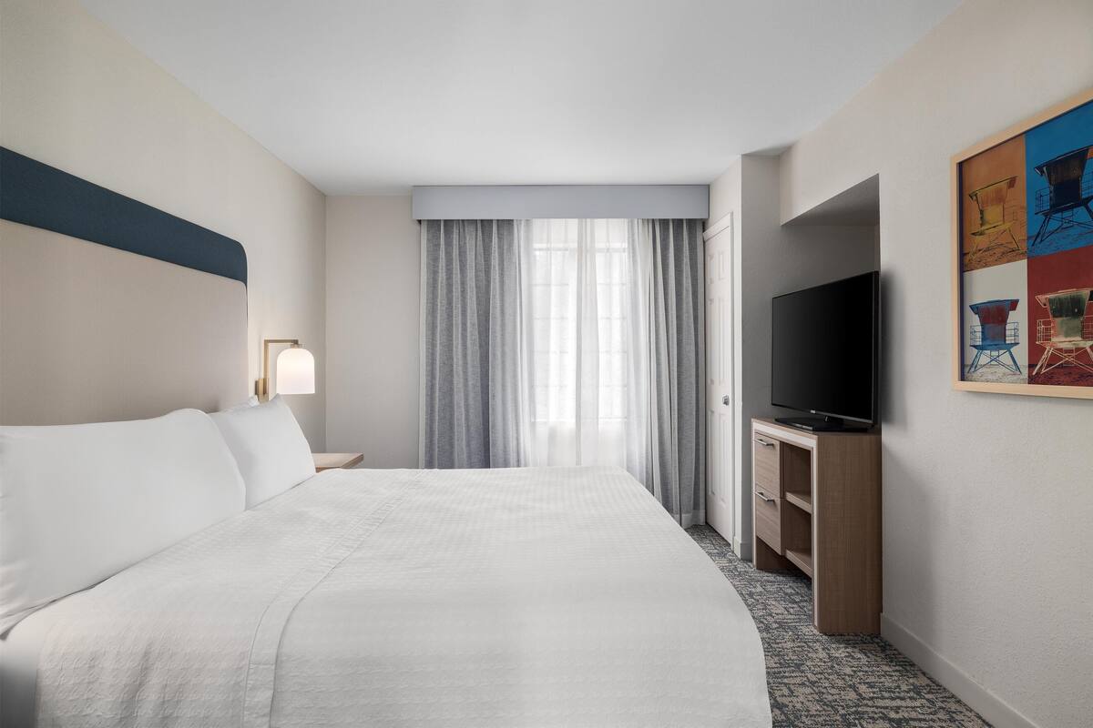 Quarto de hotel com uma cama grande, uma TV em um suporte de madeira, um quadro na parede e uma janela com cortinas.