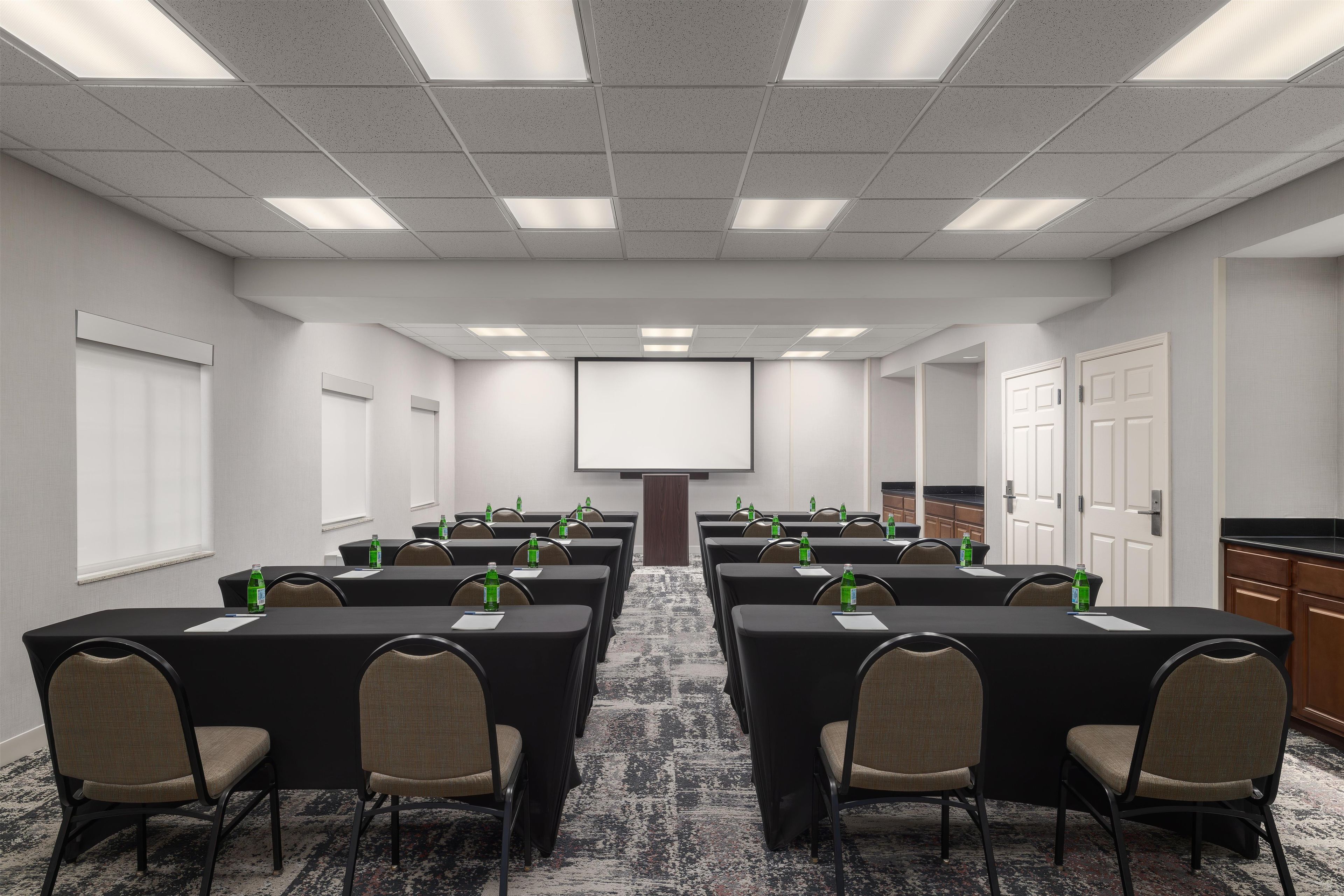Un salón de conferencias con mesas, sillas, una pantalla para proyector y botellas de agua.