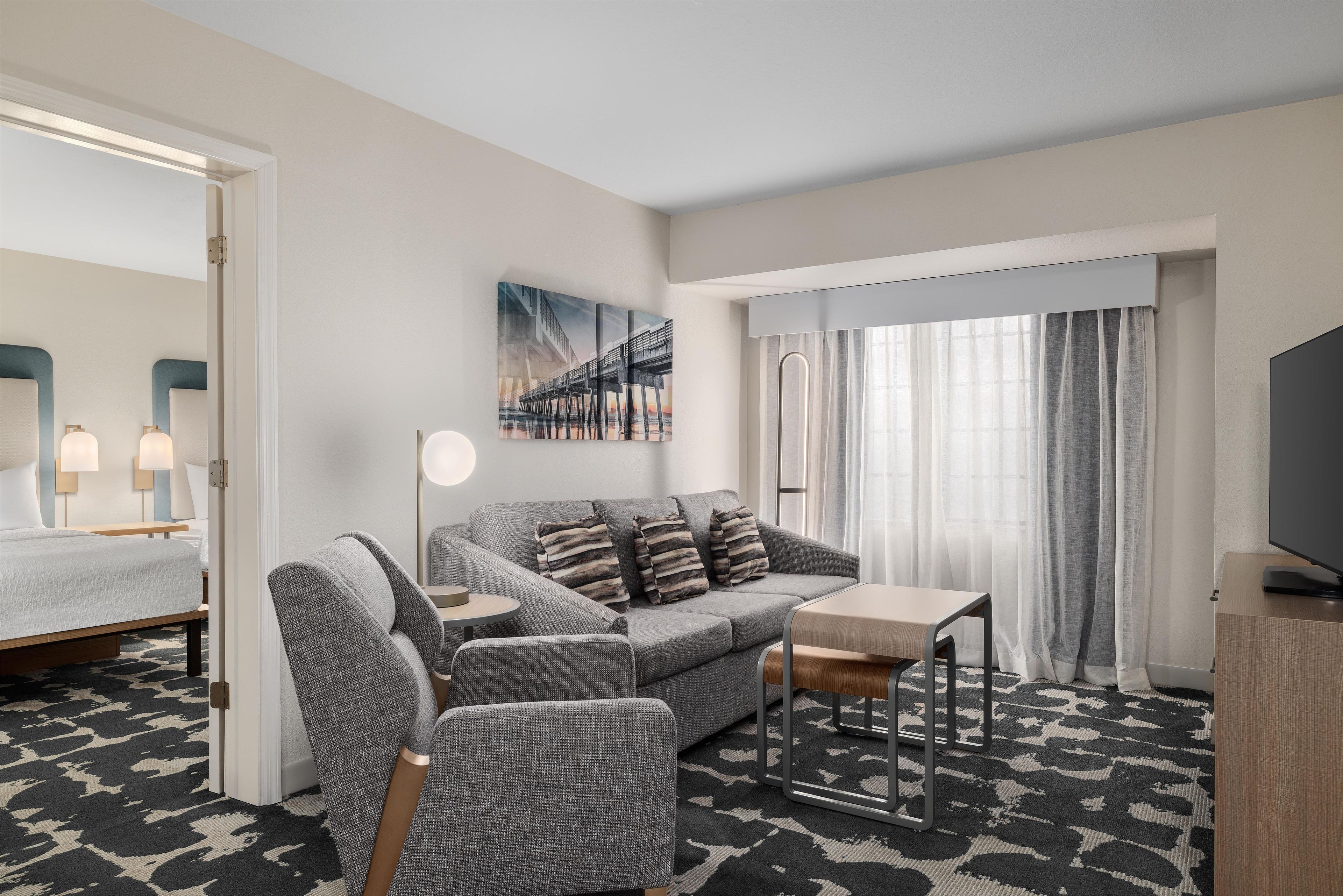 A modern hotel room with a grey sofa, a small table, a chair, a bed, a TV, and a painting on the wall.