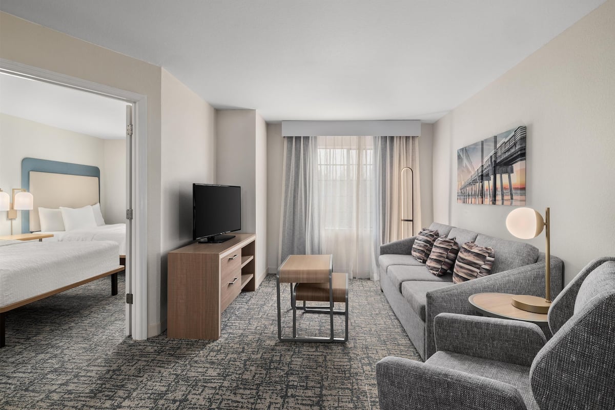 Quarto de hotel com cama, sofá, televisão, mesa pequena e um quadro na parede.