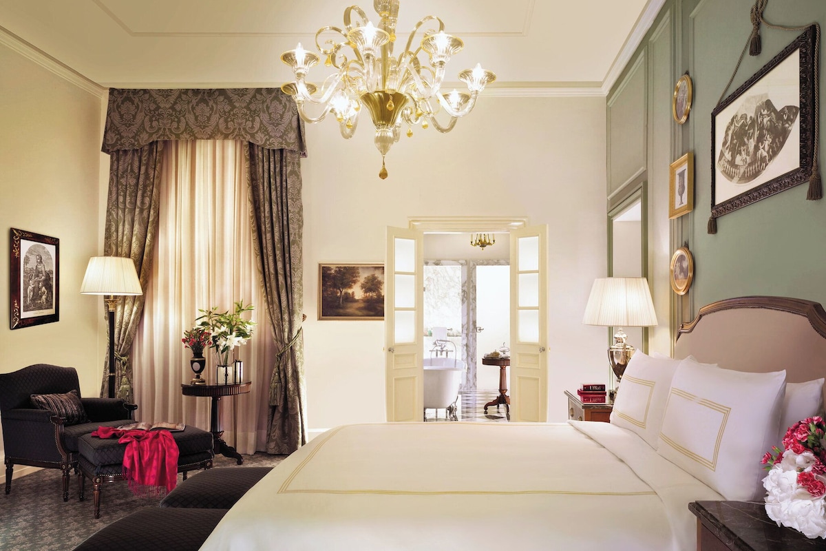 A hotel room with a large bed, a sitting area with a chair and a table, a chandelier, and a view through an open door.