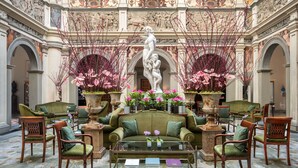 A grand lobby with a central green sofa, flanked by wooden chairs, a glass coffee table, and a marble floor. A large statue and frescoes adorn the walls.