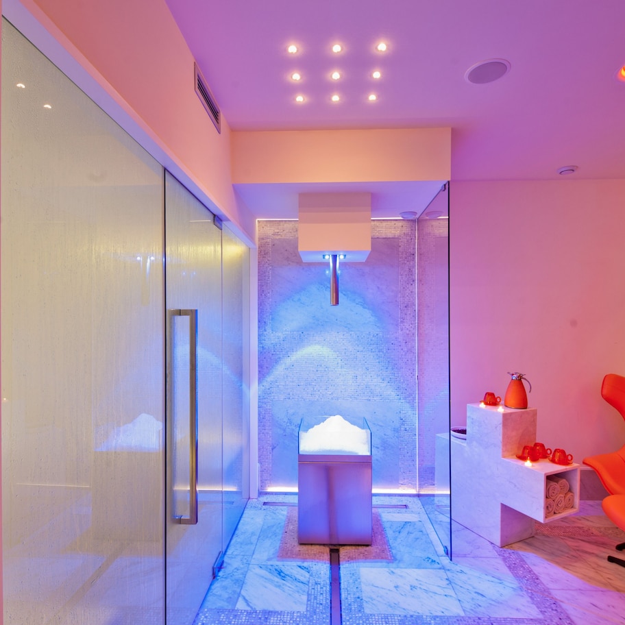 A modern spa room with a glass wall, a massage chair, a massage table, and a wall-mounted massage head.