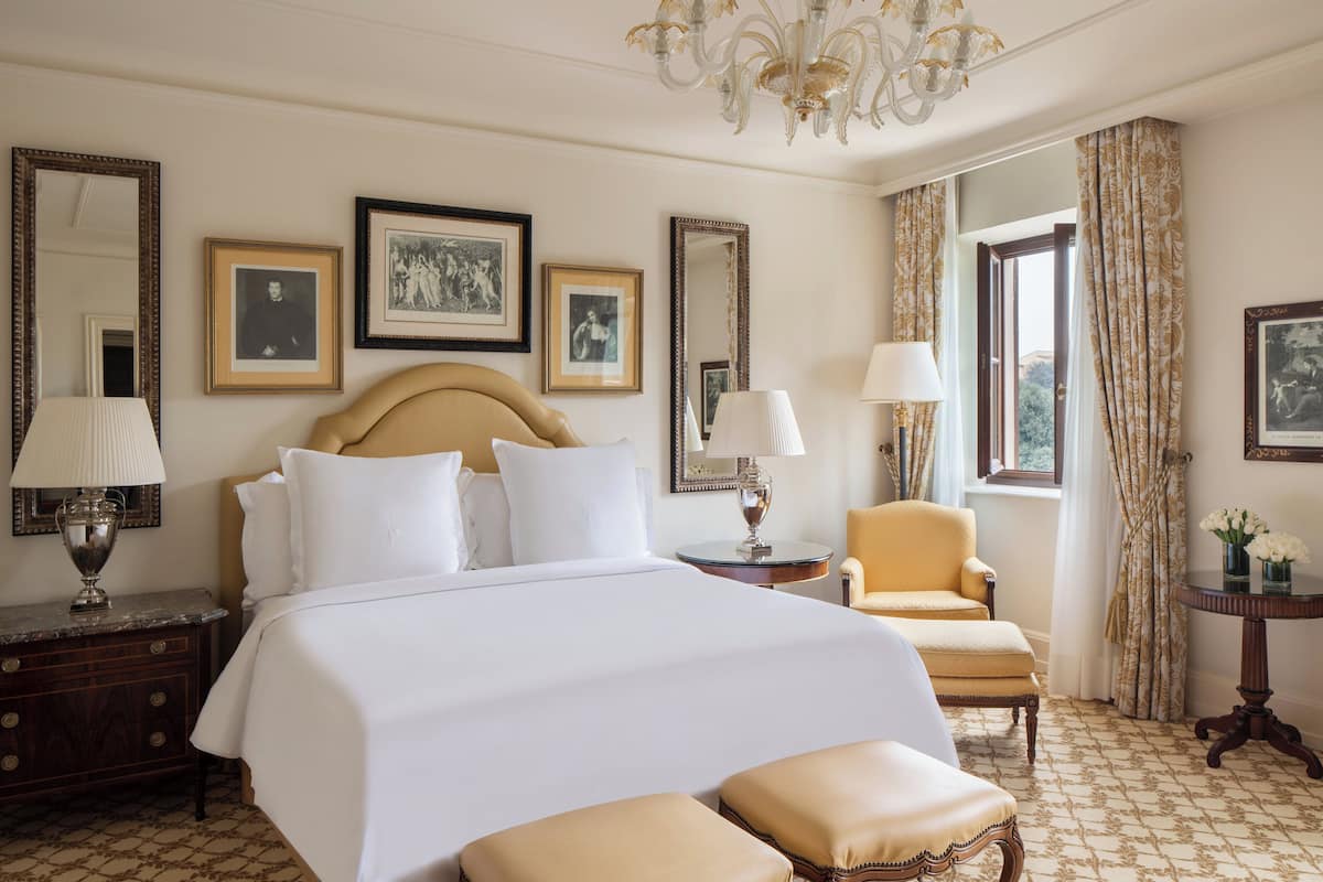 Habitación de hotel con una cama grande, dos mesitas de noche, una silla, una pequeña mesa con flores y una ventana con cortinas.