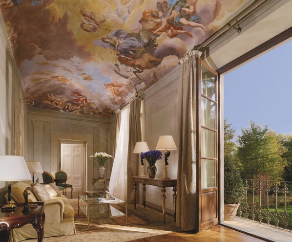 A luxurious room with a grand ceiling fresco, elegant furniture, and a large window offering a view of the outdoors.