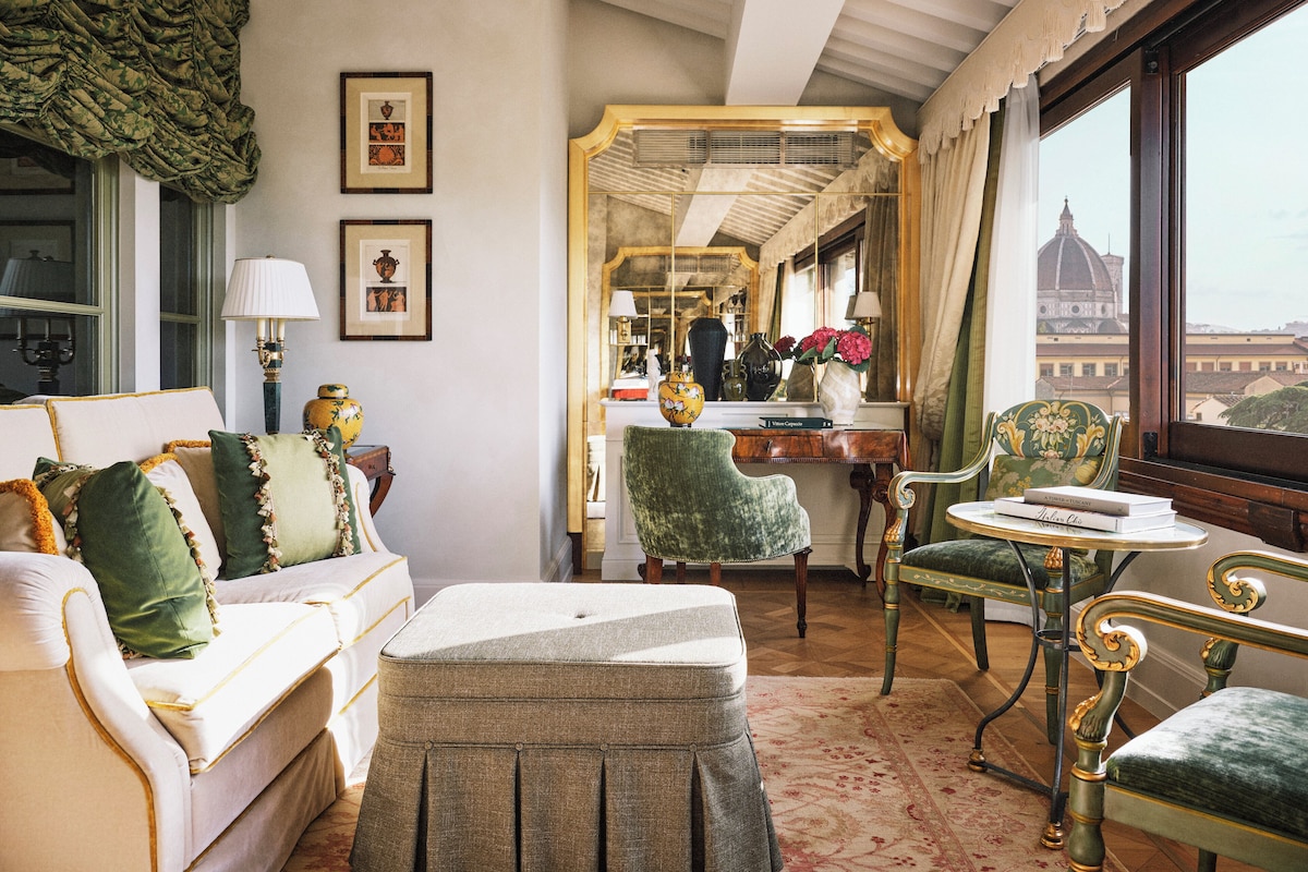 A room with a sofa, ottoman, chairs, a table, and a mirror. There are framed pictures on the wall and a window with a view of a building.