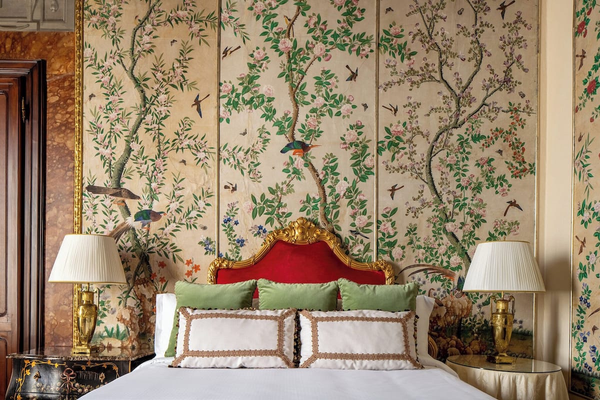 Un dormitorio con papel tapiz floral, un cabecero tapizado en rojo y dos lámparas de noche.