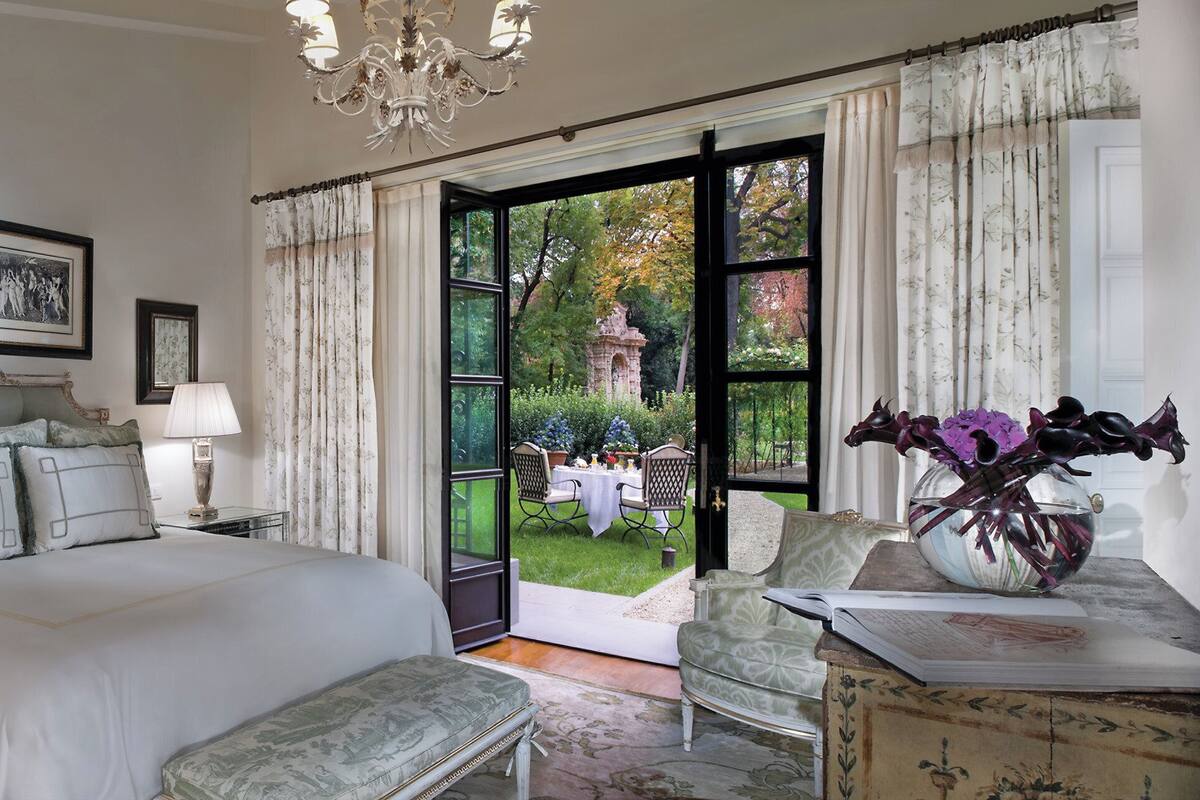 Un dormitorio con una cama grande, dos mesitas de noche con lámparas, una araña, una silla, un jarrón con flores y vistas al jardín a través de una puerta abierta.