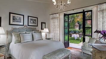 A bedroom with a large bed, two bedside tables with lamps, a chandelier, a chair, a vase with flowers, and a view of the garden through an open door.