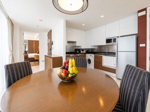 Habitación de hotel moderna con comedor que incluye una mesa de madera y sillas a rayas, cocina con muebles blancos y electrodomésticos de acero inoxidable, y un ventanal con cortinas.
