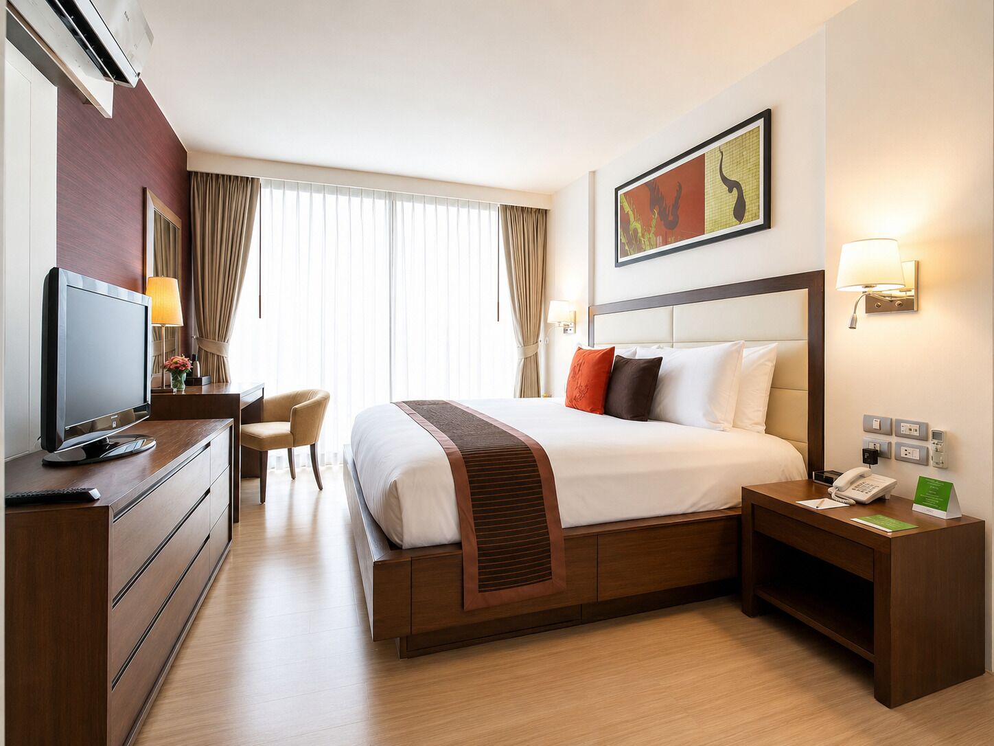A hotel room with a bed, a desk with a computer, a chair, a TV, a bedside table with a lamp, and a painting on the wall.