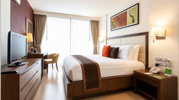 A hotel room with a bed, a desk with a computer, a chair, a TV, a bedside table with a lamp, and a painting on the wall.