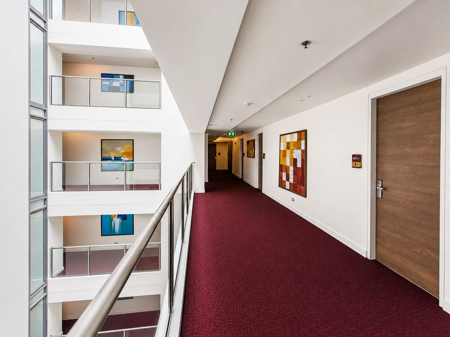 A multi-level hotel corridor with a red carpet, glass railings, and artwork on the walls.