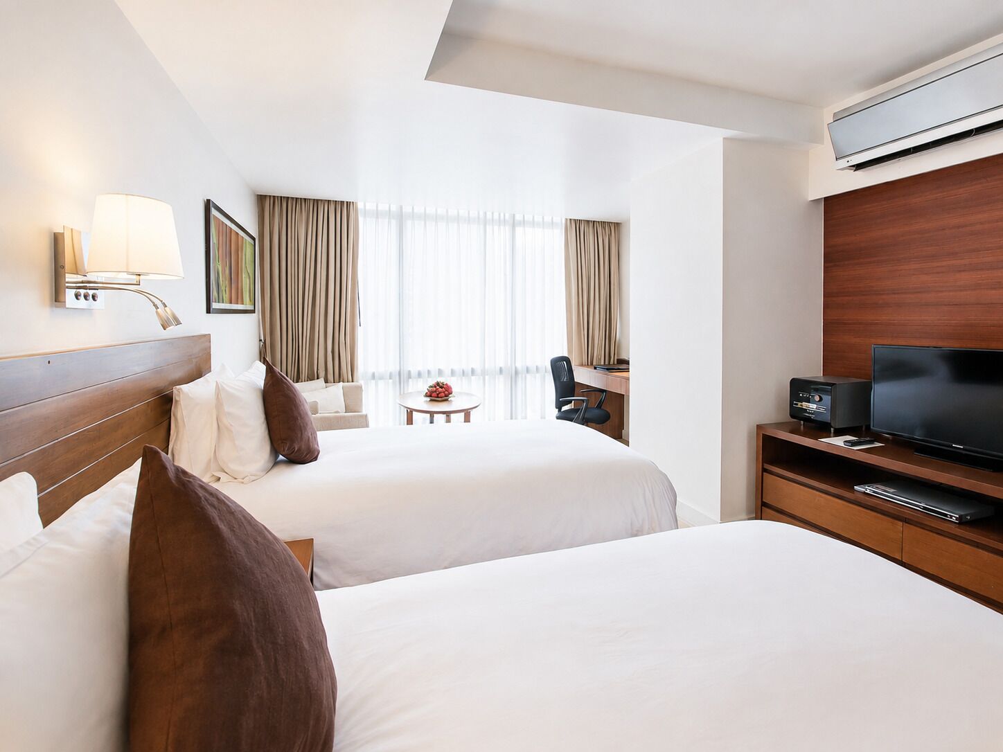 Une chambre d’hôtel avec deux lits, un bureau avec une télévision, une lampe et une fenêtre avec des rideaux.