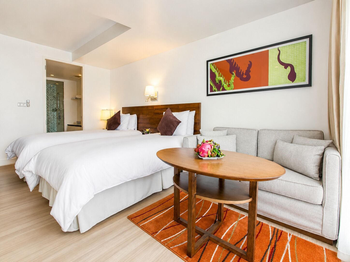 A hotel room with a bed, a sofa, a table with fruit, a painting, and a shower area.