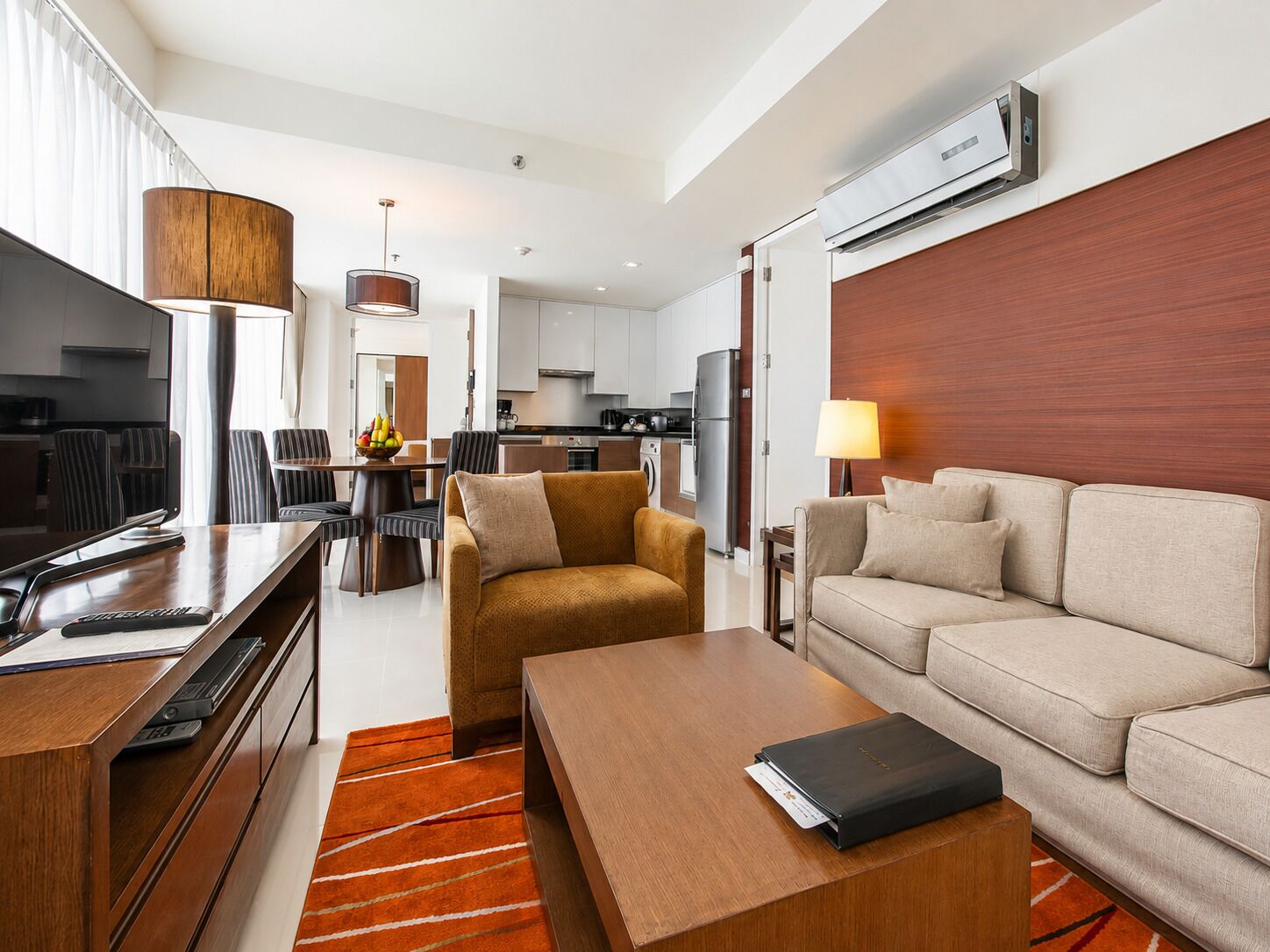 A modern hotel room with a sofa, a coffee table, a TV, a dining area, and a kitchen.