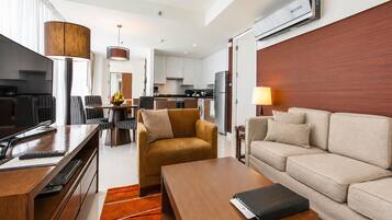 A modern hotel room with a sofa, a coffee table, a TV, a dining area, and a kitchen.