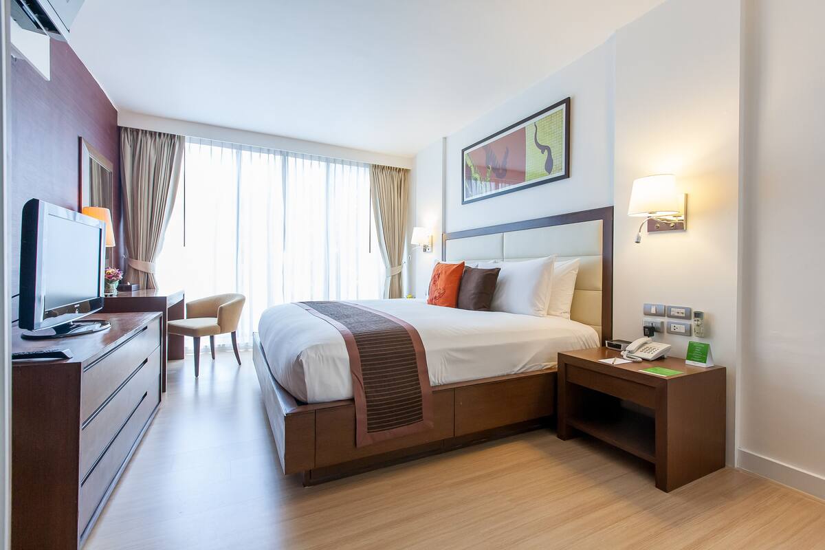 A hotel room with a bed, a desk with a computer, a chair, a bedside table with a phone, and a wall-mounted lamp.