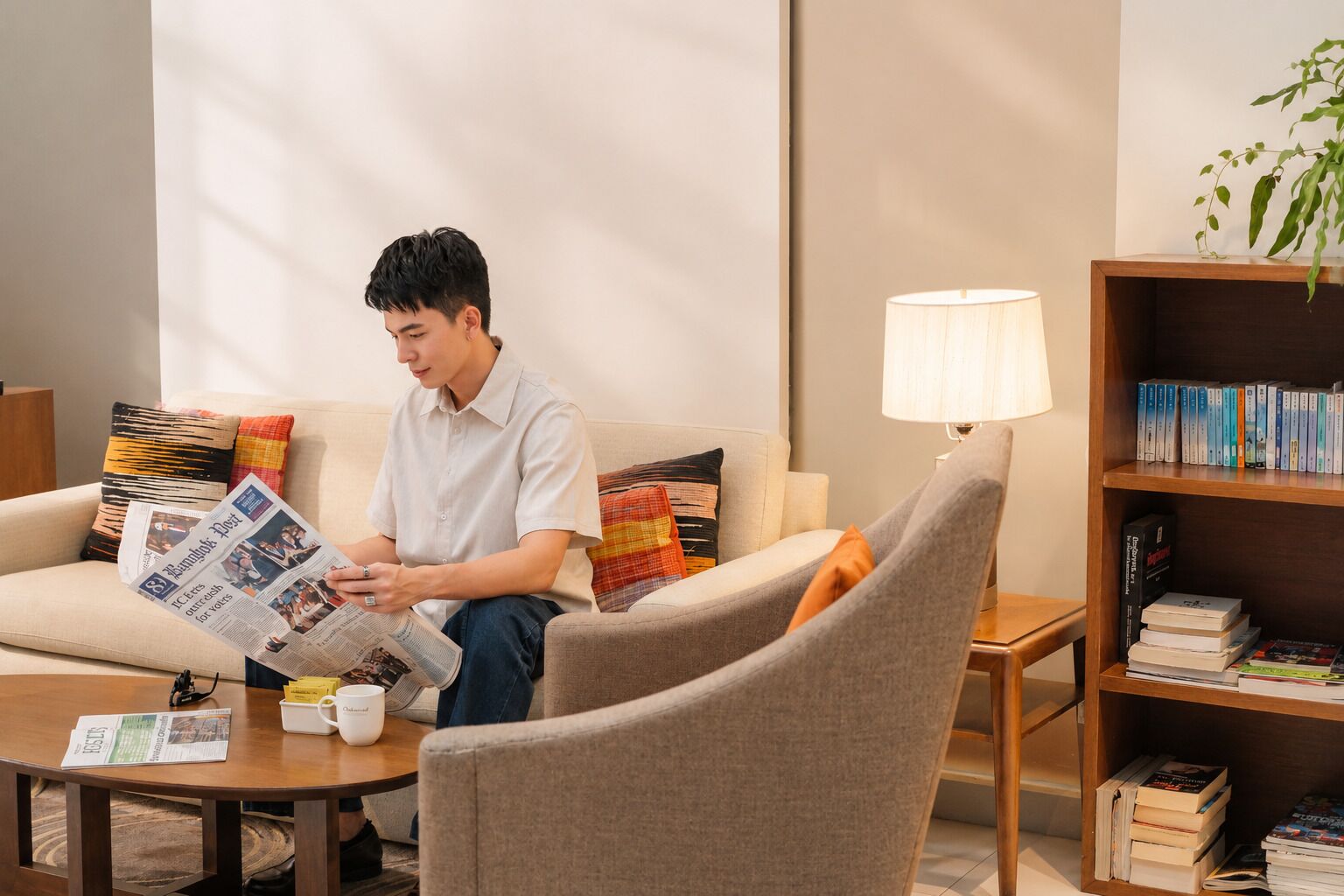A person sitting on a sofa reading a newspaper, with a coffee cup and a lamp on a side table, and a bookshelf with books.