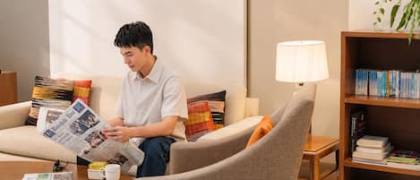 Une personne assise sur un canapé, lisant un journal, avec une tasse de café et une lampe sur une table d’appoint, et une bibliothèque remplie de livres.