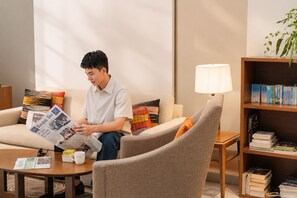 A person sitting on a sofa reading a newspaper, with a coffee cup and a lamp on a side table, and a bookshelf with books. - Oakwood Residence Sukhumvit 24 Bangkok (Bangkok)