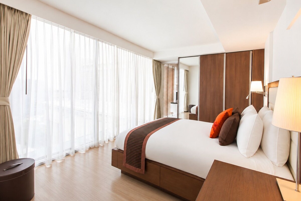 Habitación de hotel moderna con una cama grande, una mesita de noche, una lámpara y vistas a la ciudad a través de amplios ventanales.