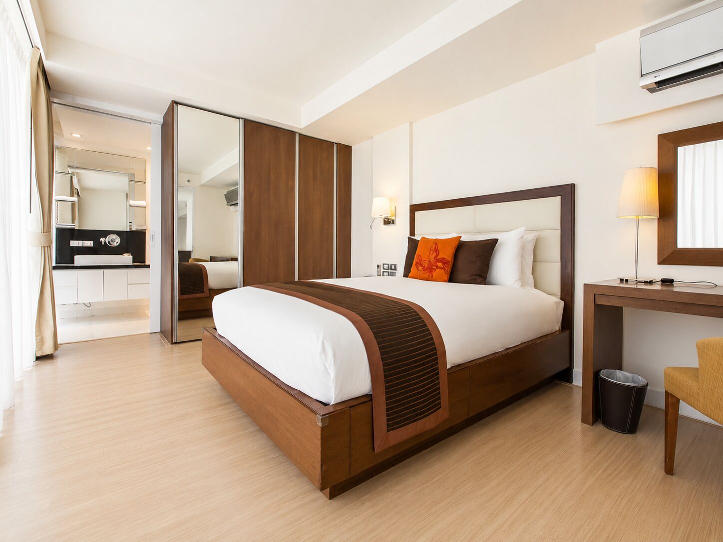 A modern hotel room with a large bed, a desk, a chair, a trash bin, and a bathroom visible through an open door.