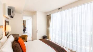Ein Hotelzimmer mit einem großen Bett, einem Schreibtisch, einem Sessel und Blick auf die Stadt durch transparente Vorhänge.