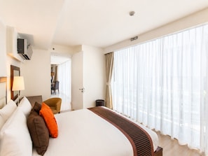 A hotel room with a large bed, a desk, a chair, and a view of the city through sheer curtains. - Oakwood Residence Sukhumvit 24 Bangkok (Bangkok)