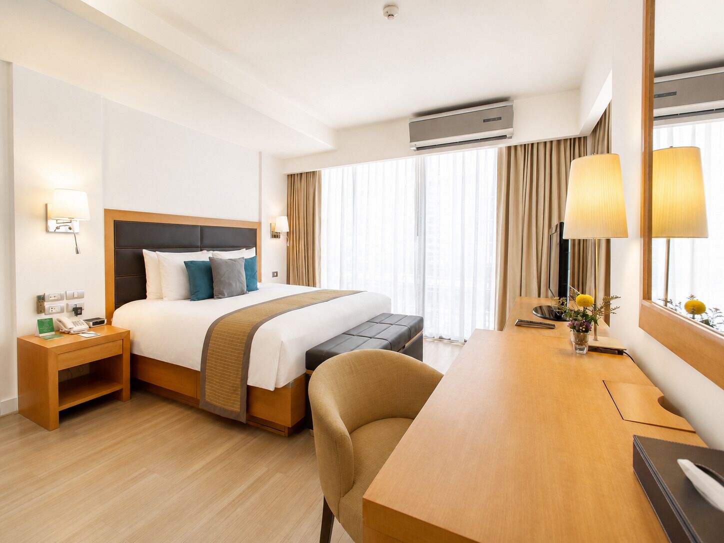 A hotel room with a bed, bedside table, lamp, desk, chair, and an air conditioning unit.