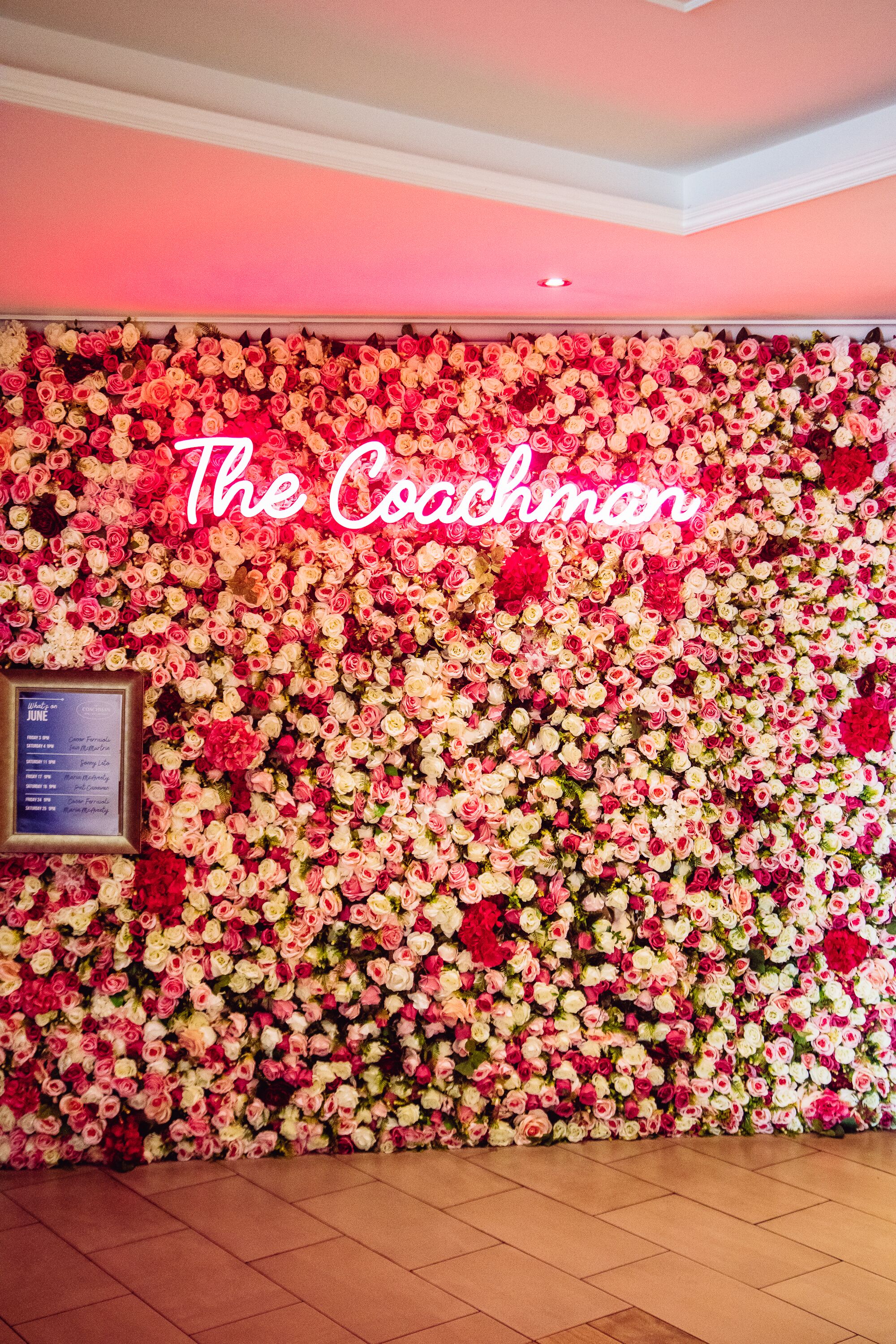 Una pared cubierta por una densa disposición de flores rosadas y blancas, con un letrero de neón que dice "The Coachman".