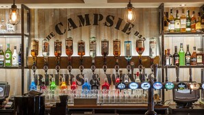 A bar area with a wooden backdrop displaying the name 'The Campsie Bar'. There are multiple liquor bottles and a variety of beer taps. - Coachman Hotel (Glasgow)