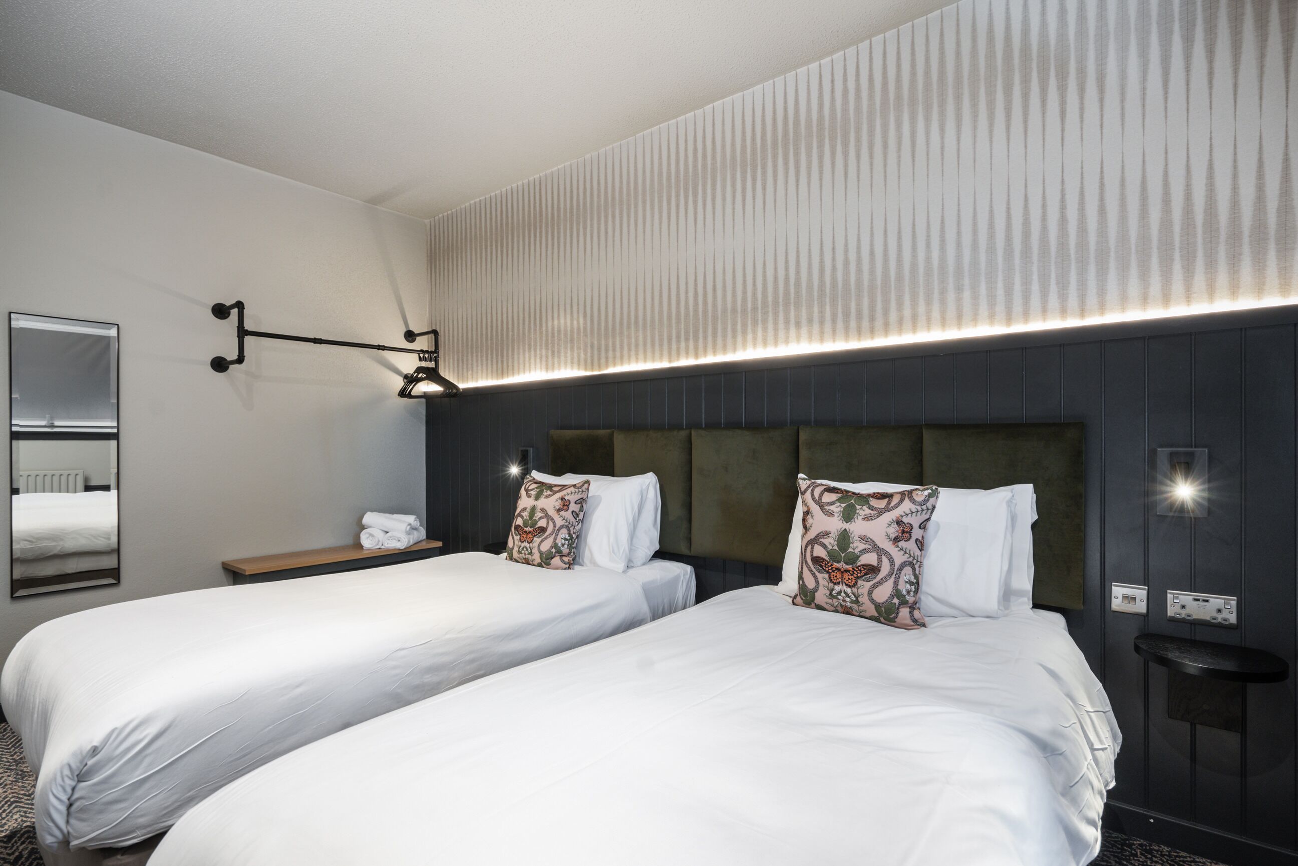 Habitación de hotel con dos camas, una mesita de noche, un espejo y una lámpara fijada en la pared.