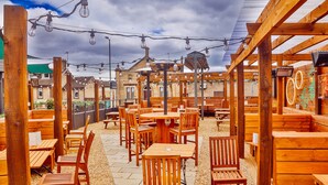 An outdoor patio with wooden tables and chairs, string lights, and a pergola structure. - Coachman Hotel (Glasgow)