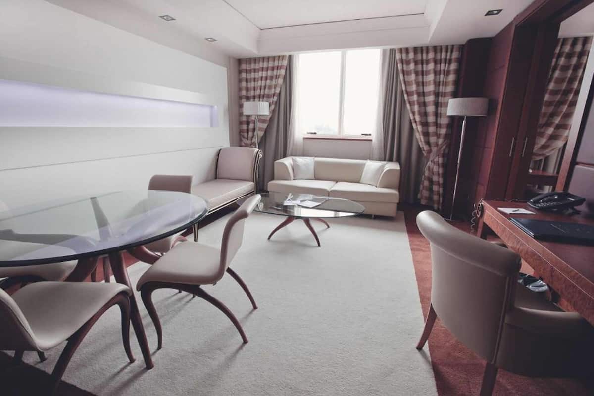 A modern hotel room with a glass table, two sofas, a desk with a lamp, and a large window with curtains.