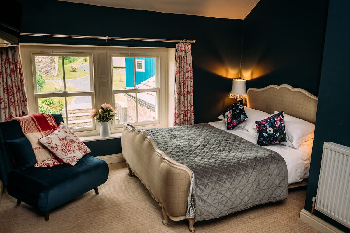 A bedroom with a bed, a chair, a window with curtains, and a nightstand with a lamp.