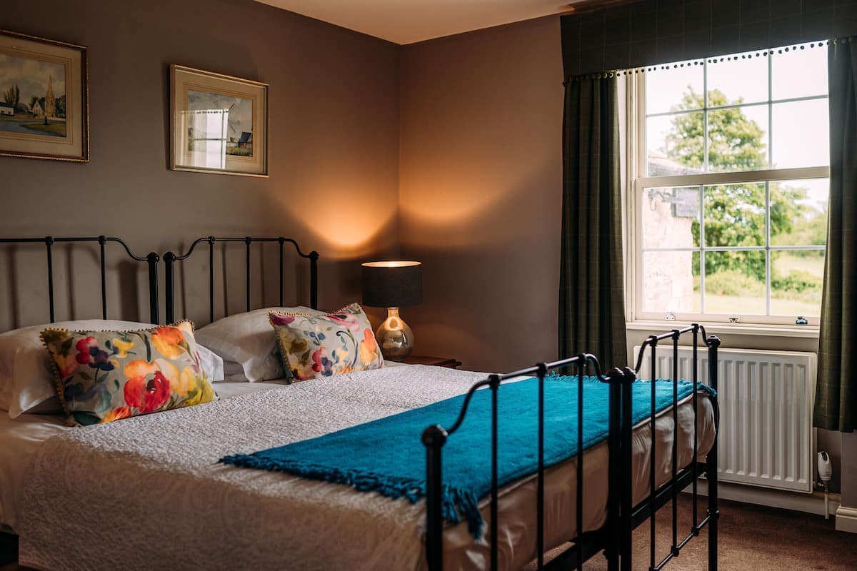 A bedroom with a bed, a nightstand, a lamp, a window with curtains, and two framed pictures on the wall.