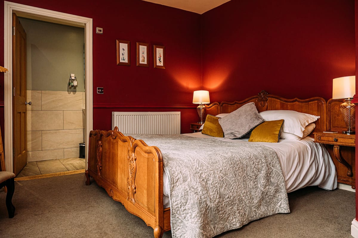 A bedroom with a wooden bed, two bedside lamps, a radiator, and a door leading to a bathroom.