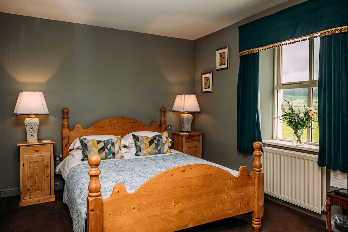 A bedroom with a wooden bed, two bedside tables with lamps, a window with curtains, and a radiator.
