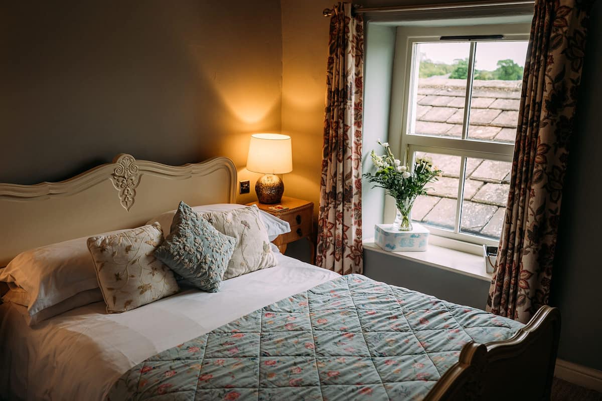A neatly made bed with floral bedding, a bedside lamp, and a window with curtains.