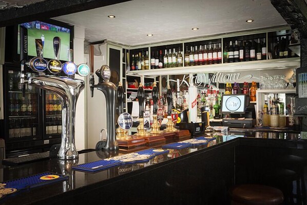A bar area with multiple taps, shelves of bottles, and a television screen displaying a sports event. - King William (Luton)