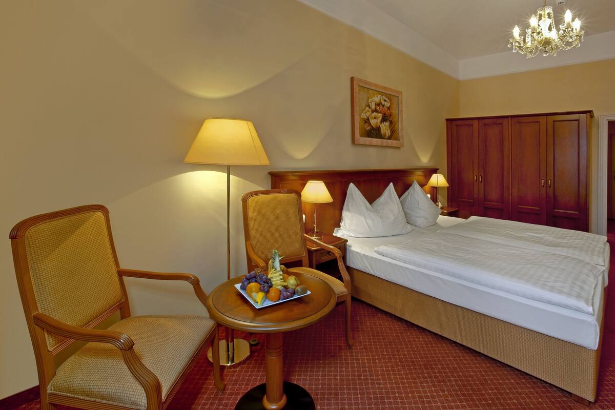 A hotel room with a bed, two chairs, a small table with fruit, and a painting on the wall.