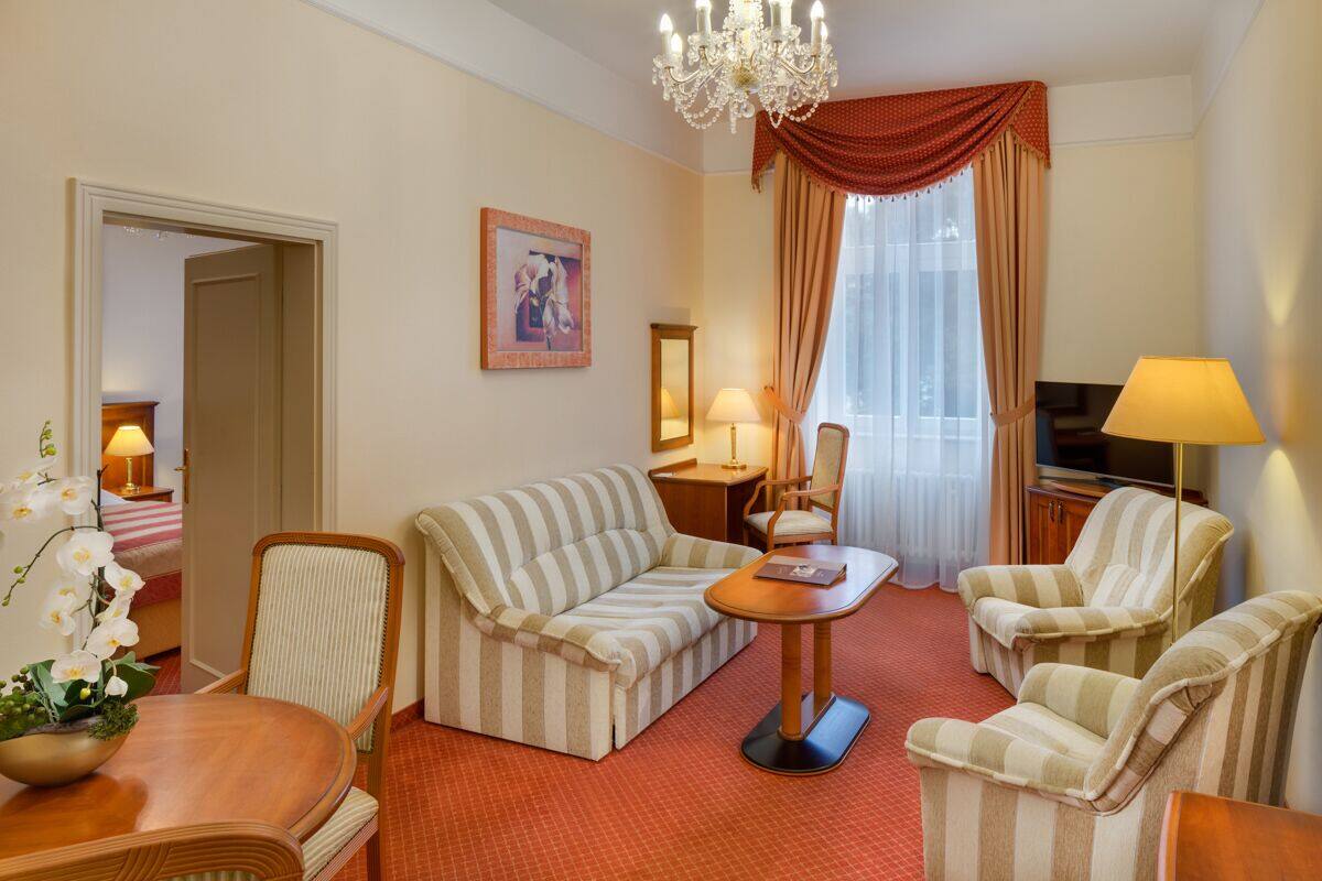 A hotel room with a striped sofa, two armchairs, a wooden coffee table, a TV, and a chandelier.