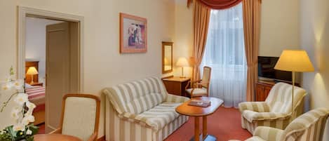 A hotel room with a striped sofa, two armchairs, a wooden coffee table, a TV, and a chandelier.