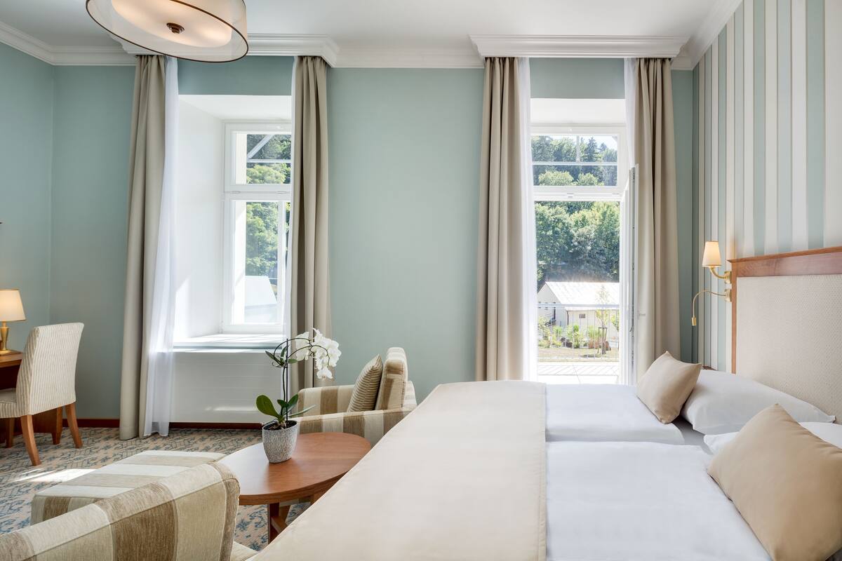 A hotel room with a large bed, a seating area with a table and chairs, a potted plant, and a view of greenery outside.