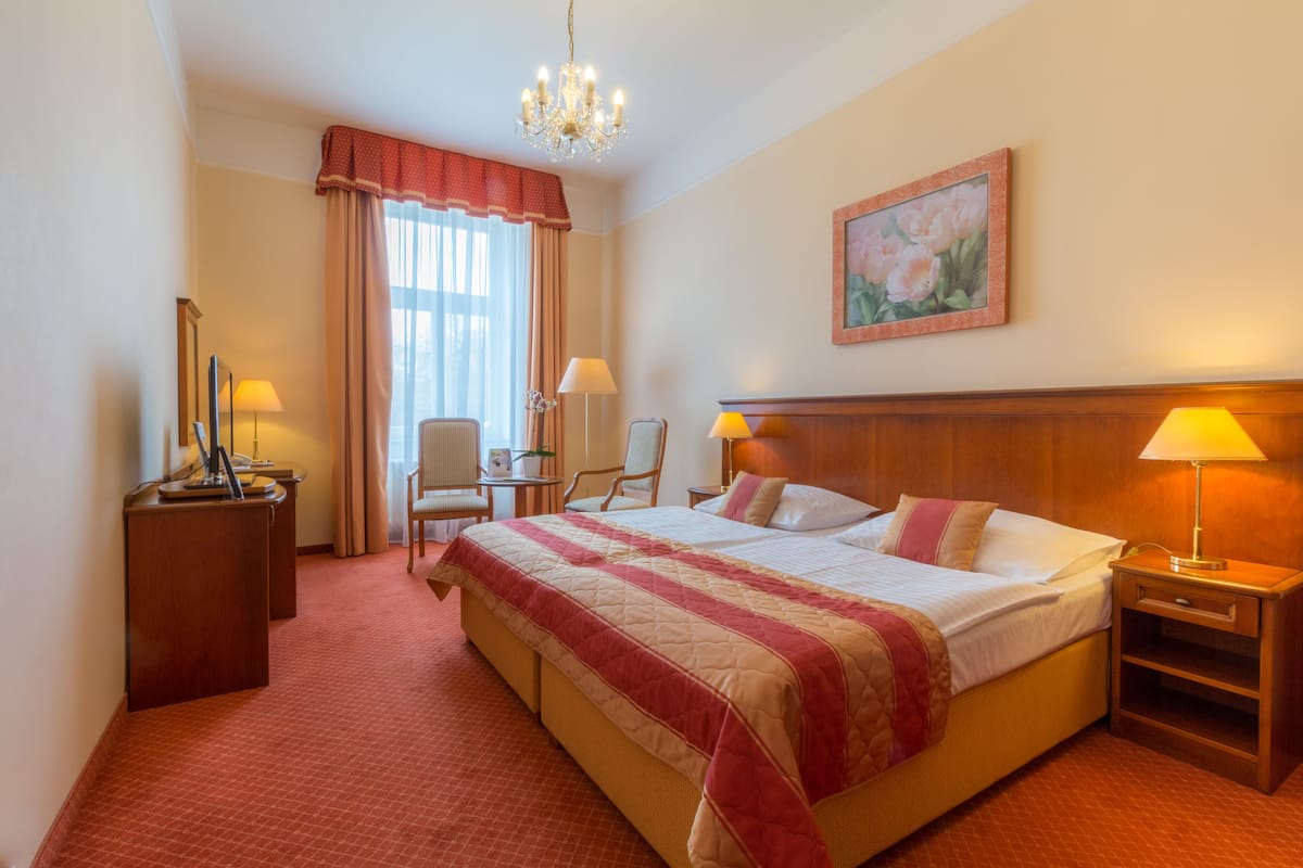 A hotel room with a large bed, two bedside tables, a chair, a desk, a chandelier, and a painting on the wall.